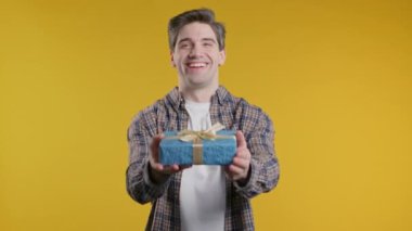 Heyecanlı Amerikalı adam hediye kutusunu tutuyor ve Sarı Duvar arkaplanındaki kameraya elleriyle veriyor. Gülümseyen adam, hediyeyle mutlu. Stüdyo resmi.