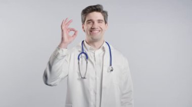 Beyaz önlük formundaki 30 'lu yaşlarda pozitif bir doktor el işaretini onaylıyor. Mutlu adam, doğru seçim, harika bir anlaşma, stüdyo geçmişi. Yüksek kalite 4k