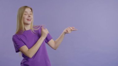 Gülümseyen sarışın bayan reklamınız için boş bir yer arıyor. Önerilen kadın sağ tarafı işaret ediyor, boşluğu kopyalayın. Kadın, menekşe arkaplanda izole bir şey sunuyor. Yüksek kalite 4k