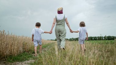 Genç bir aile, annem ve iki erkek kardeşim buğday tarlası yakınlarındaki yeşil yolda yürüyorlar. Mutlu tanınamaz anne ve çocuklar, aşk, özgürlük, gelecek, ev, kırsal, yeşil çocukluk