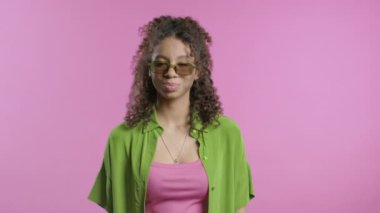 Pembe stüdyo arka planında kameraya gülümseyen Afro-Amerikan gözlüklü şık genç kız. Yeşil tişörtlü, yaz tatili kıyafetli hoş bir bayan. Yüksek kalite 4k
