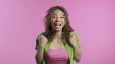 Güzel, Afro-Amerikan bir kadın zafer işareti gösteriyor. Evet, başarıyor. Hedeflerine ulaşıyor. Mutlu, mutlu, şaşırmış pembe arka plandaki mutlu bayan. Büyük ikramiye. Yüksek kalite 4k