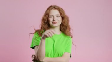 20 'li yaşlarda, neşeli, kızıl saçlı, gülümseyen, pembe stüdyo arka planında kameraya flört eden bir kadın. Yeşil elbiseli sevimli, neşeli bayan. Yüksek kalite 4k