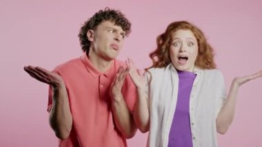 Kafası karışık, şık çift omuzlu. Yardımcı olamıyor, bilmemek için jest yapıyor. Zor soru, suçlu tepkisi, pembe arka plandaki şaşkın adam ve kadın. Yüksek kalite 4k