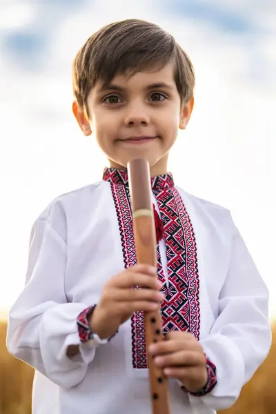 stock image Portrait of little boy child with woodwind wooden flute - ukrainian sopilka, tylynka on sky background. Folk music concept. Musical instrument. Kid in traditional embroidered shirt - Vyshyvanka.