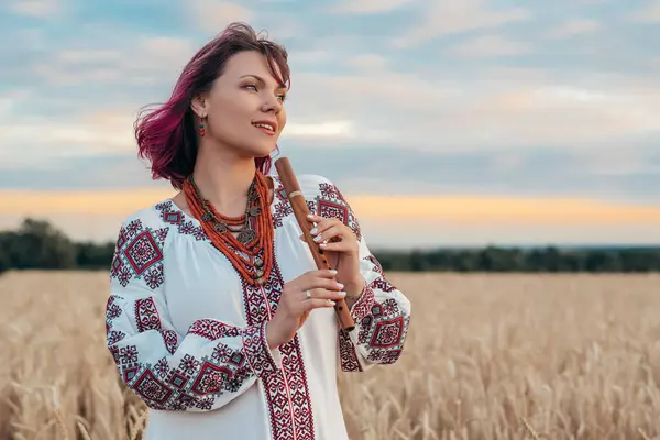 stock image Portrait of woman with woodwind wooden flute - ukrainian sopilka in wheat field. Folk music concept. Musical instrument. Lady in traditional embroidered shirt - Vyshyvanka. High quality photo