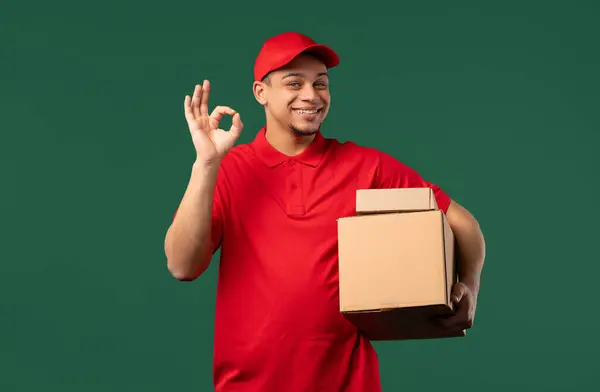 stock image Positive postal courier man with hands sign OK, okay. Happy postman in red uniform, correct perfect choice, great deal, green studio background. High quality