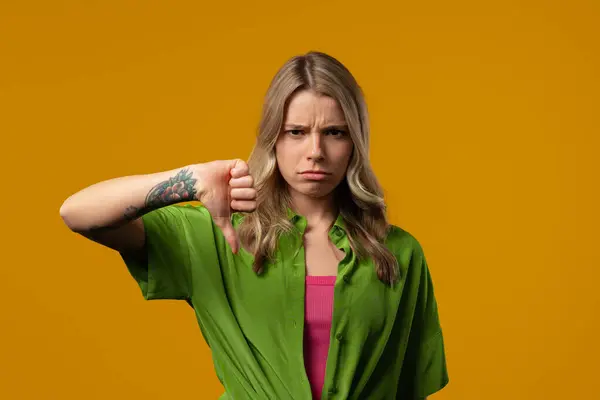 stock image Portrait of unhappy european woman condemns with sign of dislike. Young millennial lady expressing discontent with showing thumbs-down gesture on yellow studio background.