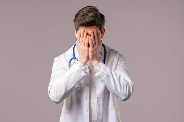 Disappointed physician man in white medical coat form doing facepalm. Feeling regret, failure, dissatisfaction, wasted time, problems in clinic, white studio background. High quality