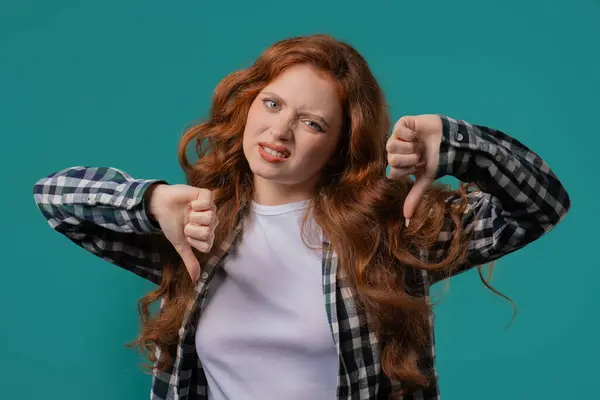 stock image Portrait of unhappy european woman condemns with sign of dislike. Young millennial lady expressing discontent with showing thumbs-down gesture on blue studio background.