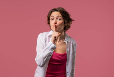 Smiling american woman holding finger on lips, pink studio background. Pretty lady with gesture of shhh, secret, silence, conspiracy, gossip concept. High quality clipart