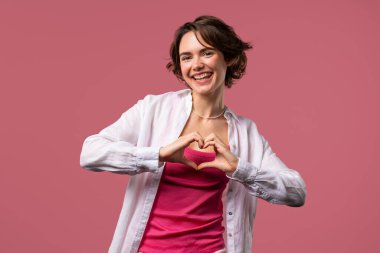 Gülümseyen güzel bir kadın, kalp şeklinde bir işaret gösteriyor, sevgisini gönderiyor. Pembe arka planda hoş bir bayan. Kadın sağlığı, gönüllülük, bağış, minnettarlık sembolü, flört konsepti. Yüksek kalite