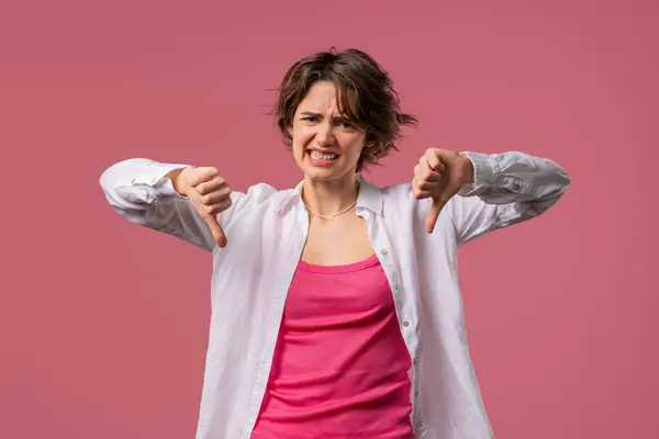 stock image Portrait of unhappy european woman condemns with sign of dislike. Young millennial lady expressing discontent with showing thumbs-down gesture on pink studio background.