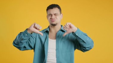 Mutsuz bir adamın portresi, hoşnutsuzluk işaretiyle kınanır. Genç milenyum çocuğu, sarı stüdyo arka planında olumsuz bir jest göstererek memnuniyetsizliğini dile getiriyor..