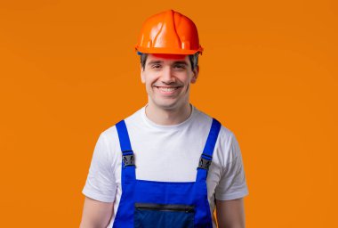 Friendly foreman in overall and helmet portrait on orange studio background. Positive smiling guy, headshot of white mature male model.  clipart