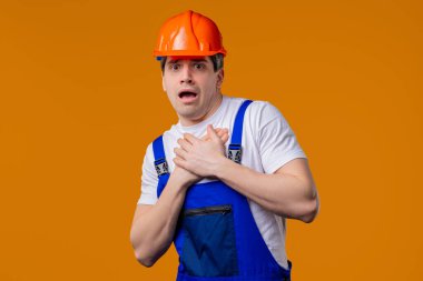 Koruyucu üniformalı ve kasklı korkmuş tamirci bir şeyden korkuyor ve turuncu arka planda korku dolu büyük gözlerle kameraya bakıyor. Fobi, sorun, panik konsepti.