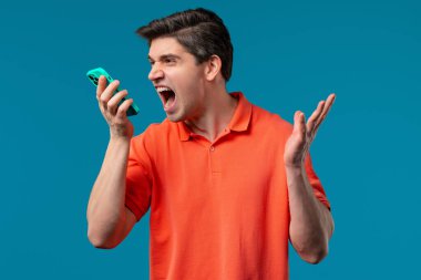 Annoyed young man quarreling while talking on mobile phone. Angry dude, stress. Blue studio background. Conflict, dispute, negative. High quality  clipart