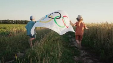 Küçük çocuklar, olimpiyatlarda koşan taraftarlar kırsal yolda rüzgarda dalgalanan bayrak sallıyor. Uluslararası spor müsabakası yaklaşıyor. Resmi amblem. Yüksek kalite 4k görüntü