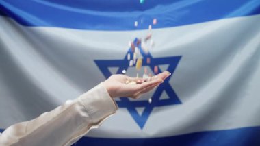 İsrail bayrağında kadın elinin üzerine düşen haplar. Pahalı ilaçlar, ilaç işi. Eczane temalı. İsrail 'de çok renkli uyuşturucu ve kapsüller.