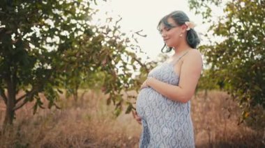 Bebek bekleyen mutlu anne karnında çocuk taşıyor. İnanılmaz annelik. Ceviz ağaçlarının arka planında güzel elbiseli hamile kadın, kırsal kesimde. Yüksek kalite 4k görüntü