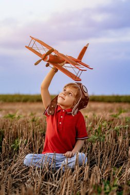 Gülümseyen 5 yaşındaki çocuk tahta uçakla oynuyor, gelecekteki mesleki ilgisini keşfediyor, hayalperest çocuk uçuyor, yaz sahasında seyahat ediyor. Yaratıcı hayal, fantezi, çocukluk.