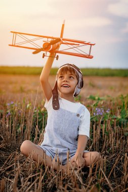 Gülümseyen 5 yaşındaki çocuk tahta uçakla oynuyor, gelecekteki mesleki ilgisini keşfediyor, hayalperest çocuk uçuyor, yaz sahasında seyahat ediyor. Yaratıcı hayal, fantezi, çocukluk.