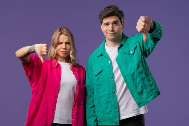 Portrait of unhappy european woman and man condemns with sign of dislike. Couple expressing discontent with showing thumbs-down gesture on violet studio background. clipart