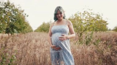 Bebek bekleyen mutlu anne karnında çocuk taşıyor. İnanılmaz annelik. Ceviz ağaçlarının arka planında güzel elbiseli hamile kadın, kırsal kesimde. Yüksek kalite 4k görüntü