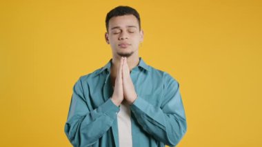 Young man praying with hands on yellow background. Guy begs someone satisfy desires, help with, prays for luck in exam. High quality 4k