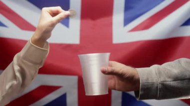 Zengin kadın, Union Jack UK bayrağında sadaka isteyen evsiz bir adamın plastik bardağına bozuk para atıyor. Paylaşmak ve birbirimize yardım etmek. Yoksul hayat, işsizlik, yoksulluk sınırının altındaki insanlar.. 