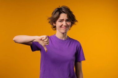 Portrait of unhappy european woman condemns with sign of dislike, shaming. Young millennial lady expressing discontent with showing thumbs-down gesture on yellow studio background. clipart