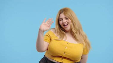Vücut pozitifliği olan genç bir kadın el sallıyor. Merhaba ya da selam ver, Chao. Ayrılıyorum, kameraya hoşça kal de. Mavi stüdyo arka planında güzel bir yaz hanımı. Yüksek kalite 4k