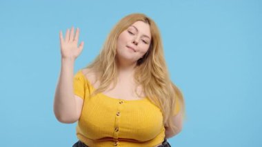 Vücut pozitifliği olan genç bir kadın el sallıyor. Merhaba ya da selam ver, Chao. Ayrılıyorum, kameraya hoşça kal de. Mavi stüdyo arka planında güzel bir yaz hanımı. Yüksek kalite 4k