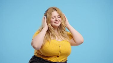 Kıvrımlı Amerikalı kadın kafayı buluyor, müzik dinliyor, mavi stüdyo arka planında kulaklıklarla dans etmekten zevk alıyor. Radyo, kablosuz modern ses teknolojisi, çevrimiçi çalar. Yüksek kalite 4k