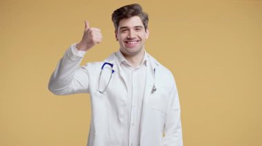 Beyaz önlük formundaki 30 'lu yaşlarda olumlu bir doktor el işaretini onaylıyor. Mutlu adam, doğru seçim, harika bir anlaşma, sarı stüdyo geçmişi. Yüksek kalite 4k