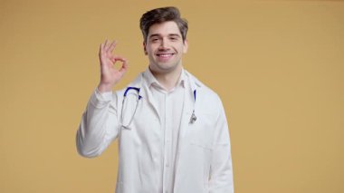 Beyaz önlük formundaki 30 'lu yaşlarda pozitif bir doktor el işaretini onaylıyor. Mutlu adam, doğru seçim, harika bir anlaşma, bej rengi sıcak stüdyo geçmişi. Yüksek kalite 4k