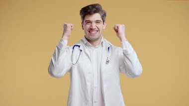 Heyecanlı beyaz önlüklü genç erkek doktor yumruğunu sıkarak muzaffer bir evet hareketi yapıyor. Tıbbi profesyoneller zaferi, başarıyı, sağlık hizmetlerinin olumlu sonucunu kutluyor. Yüksek kalite