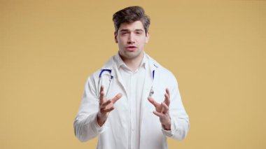 Beyaz önlüklü olgun bir erkek doktor kamerayla görüntülü konuşma yapıyor ya da video bloğu kaydediyor, tıp dersi veriyor, danışmanlık yapıyor. Bej rengi stüdyoda kendine güvenen bir sağlık uzmanı bilgisini paylaşıyor..