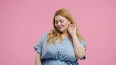 Neşeli Sarışın Artı Boyutlu Kadın Gülümseyen, Pembe Stüdyoda Kameraya Flört Eden. Çekici modayı takip eden kıvrımlı figürlü kız uzun muhteşem saçlarını düzeltiyor. Yüksek kalite 4k görüntü