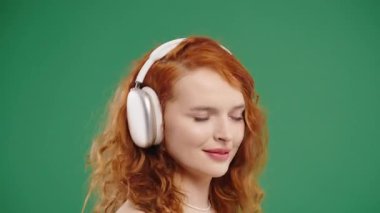 Kıvırcık Amerikalı kadın kafayı buluyor, müzik dinliyor, yeşil stüdyo arka planında kulaklıklarla dans etmekten zevk alıyor. Radyo, kablosuz modern ses teknolojisi, çevrimiçi çalar. Yüksek kalite 4k