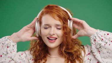 Kıvırcık Amerikalı kadın kafayı buluyor, müzik dinliyor, yeşil stüdyo arka planında kulaklıklarla dans etmekten zevk alıyor. Radyo, kablosuz modern ses teknolojisi, çevrimiçi çalar. Yüksek kalite 4k