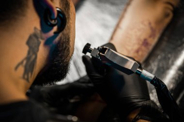 Dövmeci, müşterisi için yakın plan makine ile dövme yapıyor. Salonda dövme yaptırma işlemi. Vücut dövme işlemi. Rastalı bir adam lambanın altında kadın vücudu üzerinde çalışıyor..