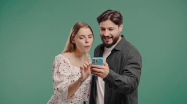 Cep telefonu aygıtı kullanan genç bir çift. Kadın ve erkek izliyor, konuşuyor, gülümsüyor, akıllı telefonla gülüyor. Sosyal uygulamalar, ağlar, teknoloji konsepti. Yeşil stüdyo geçmişi.