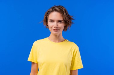 Çekici, genç bir bayan kameraya gülümsüyor, kendine güveniyor. Cazibe, bağlantı, eğlence, doğal kendini ifade etme konuları için harika. Mavi stüdyo arka planı.