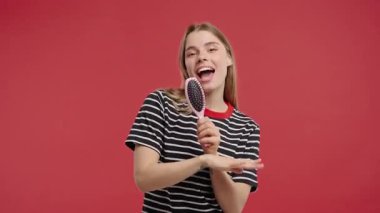 Çizgili tişörtlü neşeli sarışın kadın mikrofon gibi saç fırçasında şarkı söylüyor, parlak stüdyoda dans ediyor. Neşeli ruh hali, mutluluk, kendini ifade etme, eğlenceli sabahlar, kaygısız yaşam tarzı, kadın güveni.