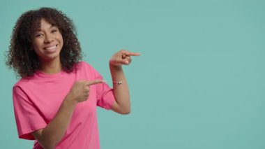 Gülümseyen öğrenci reklam için boş bir yer arıyor. Önerilen kadın fotokopi alanını işaret ediyor. Kadın, mavi arka planda izole edilmiş bir şey sunuyor. Yüksek kalite 4k