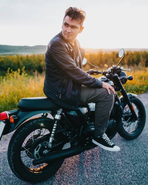 Trendy male model poses confidently on a classic black retro motorbike, showcasing urban fashion, biker lifestyle, timeless adventure vibe.