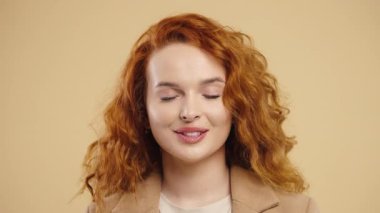 Charming young female playfully smiling at camera, confidence and light flirtation. Great for themes of charm, connection, fun, natural self-expression. Voluminous copper hair on beige wall.