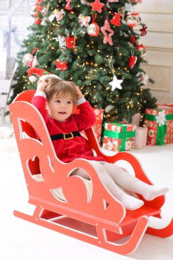 Küçük bir kız, Noel Baba gibi giyinmiş, kırmızı bir kızağın içinde, beyaz bir zeminde, bir Noel ağacının arka planında oturuyor.