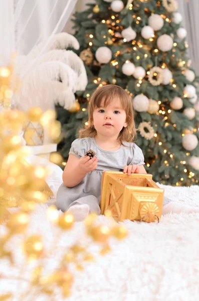一个穿着灰色连衣裙的小女孩坐在床上 背靠着一棵装饰过的圣诞树 床上有一个金黄色的礼品盒 — 图库照片